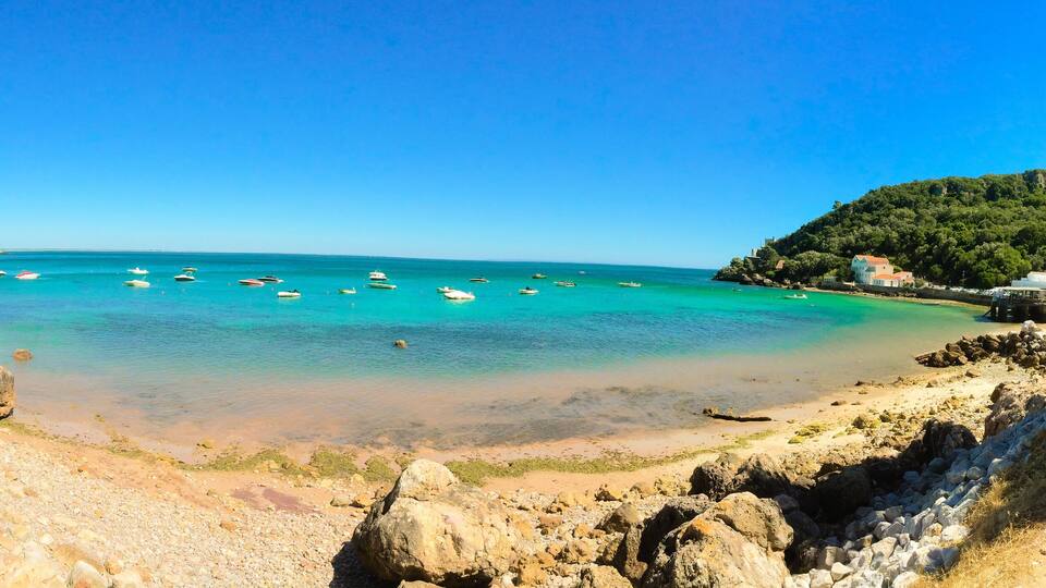 A coastal beach with clear turquoise waters, scattered rocks, and a few boats.