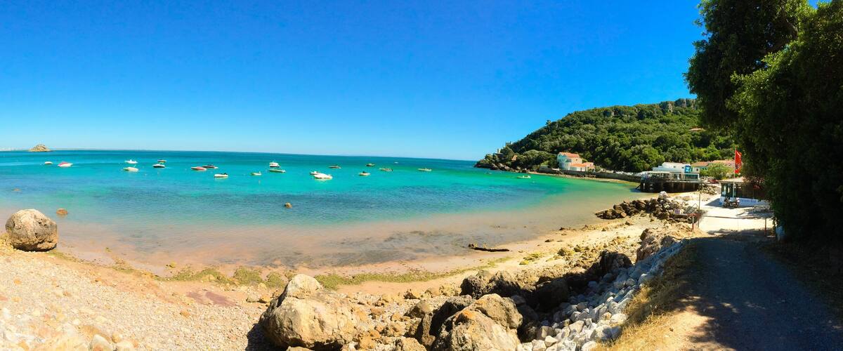 A coastal beach with clear turquoise waters, scattered rocks, and a few boats.