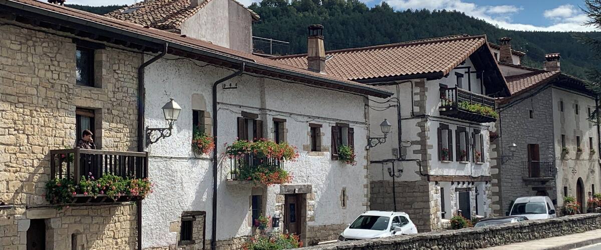 A row of stone buildings with tiled roofs, a stone wall, and a road in a mountainous area.