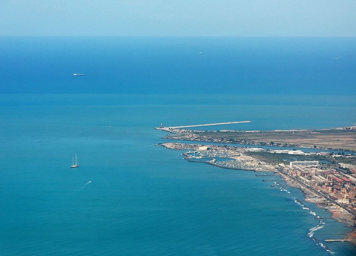 Una ciudad costera con un faro, un muelle y un velero a lo lejos.