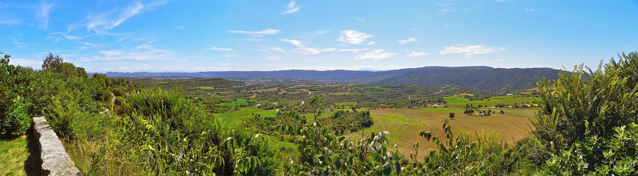 A lush valley with rolling hills, a stone wall, and distant mountains under a clear sky.