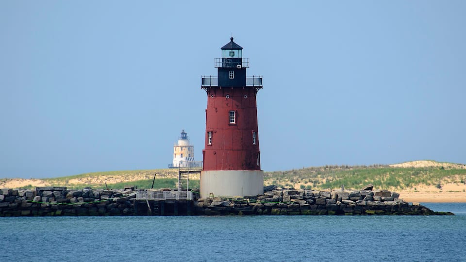 A red and white lighthouse on a rocky outcrop with calm water in the foreground and a sandy beach with dunes in the background.
