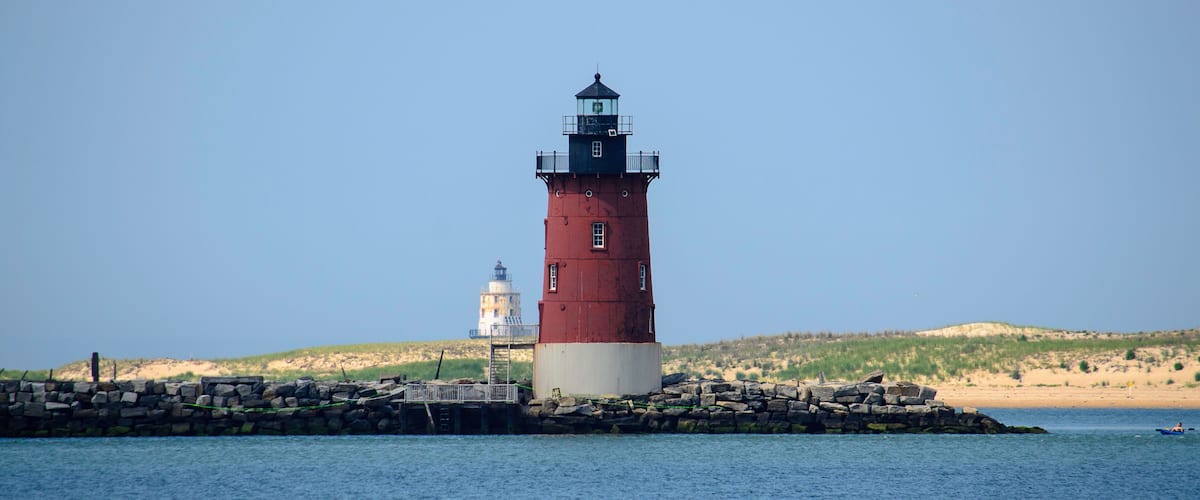 A red and white lighthouse on a rocky outcrop with calm water in the foreground and a sandy beach with dunes in the background.