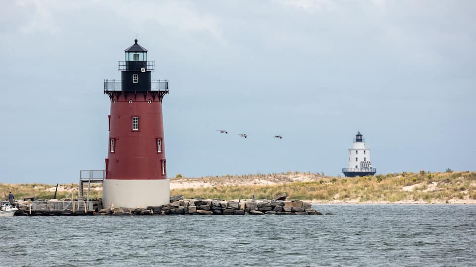 A red and white lighthouse with a smaller white lighthouse in the background, surrounded by calm water and a grassy shoreline.