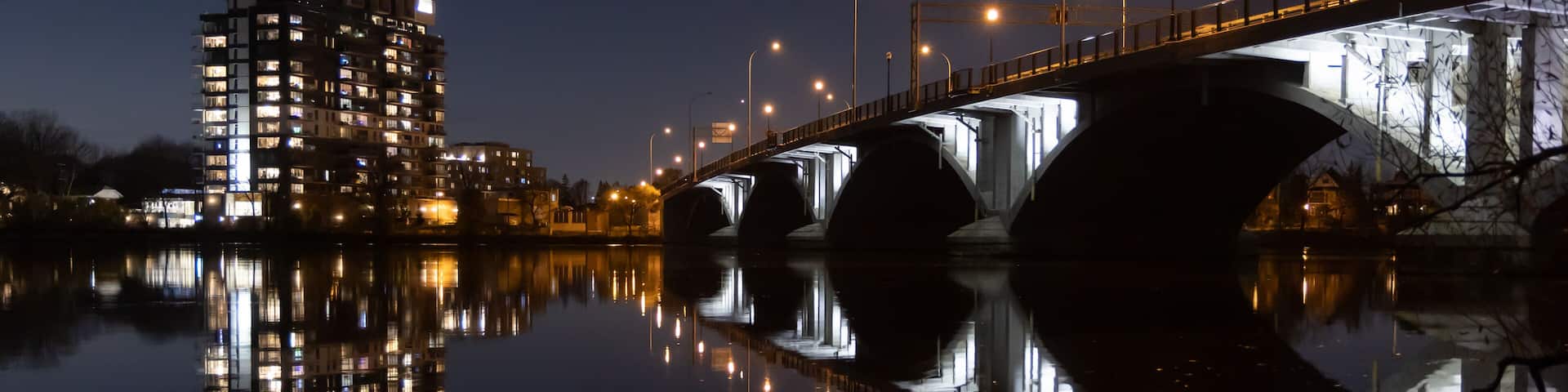 A nighttime river scene with a bridge, illuminated buildings, and their reflections on the water.