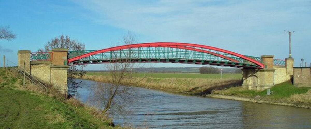 A red metal bridge over a canal with grassy banks and a clear sky.