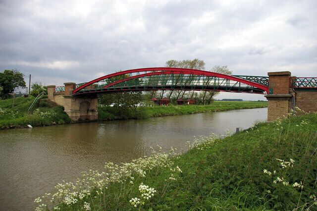 A red metal bridge over a calm river, surrounded by greenery and white flowers.