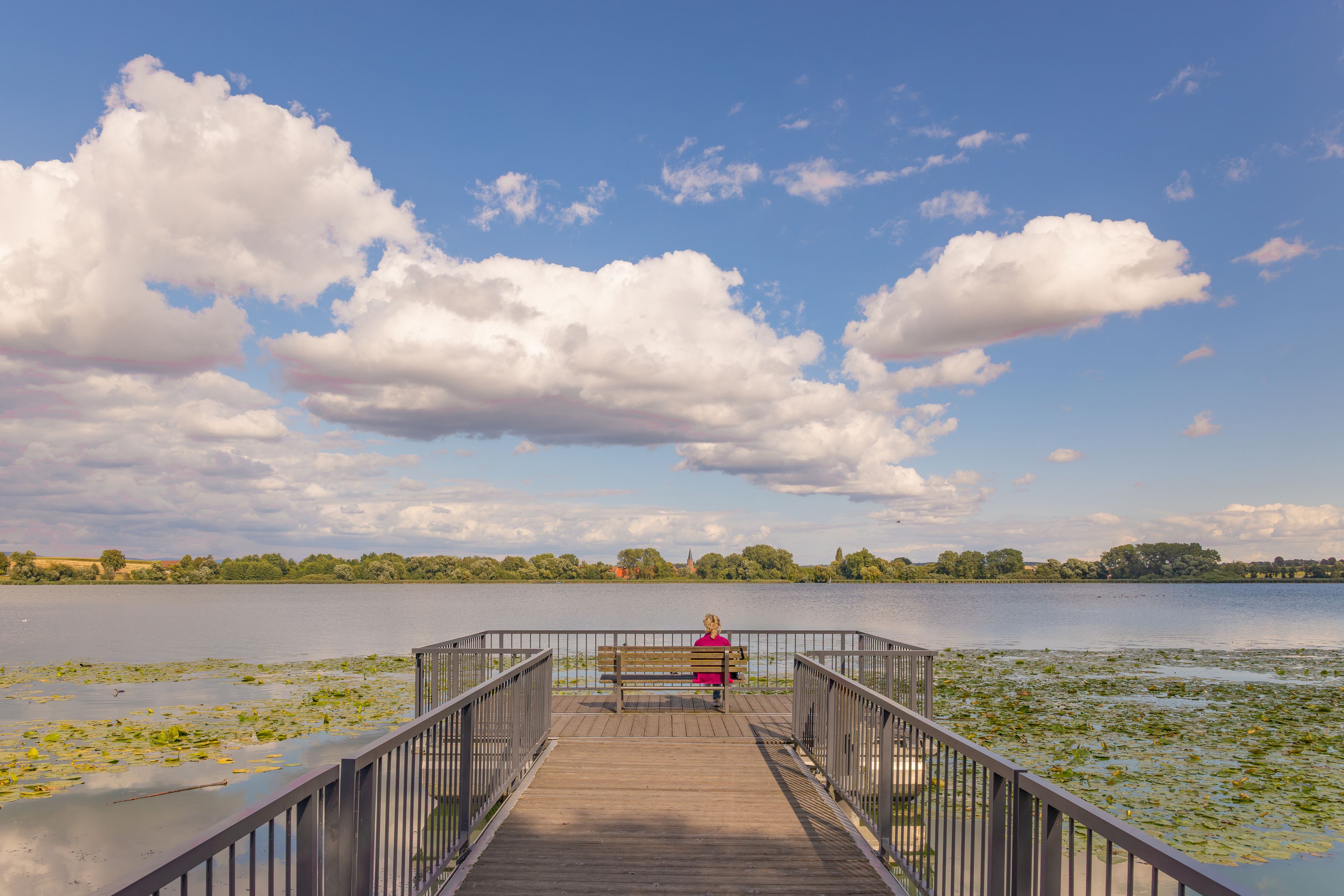 A wooden pier leading to a calm lake with lily pads, a person sitting on a bench, and a church spire in the distance.
