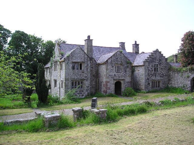 A stone building with multiple chimneys and windows, surrounded by greenery and stone walls.