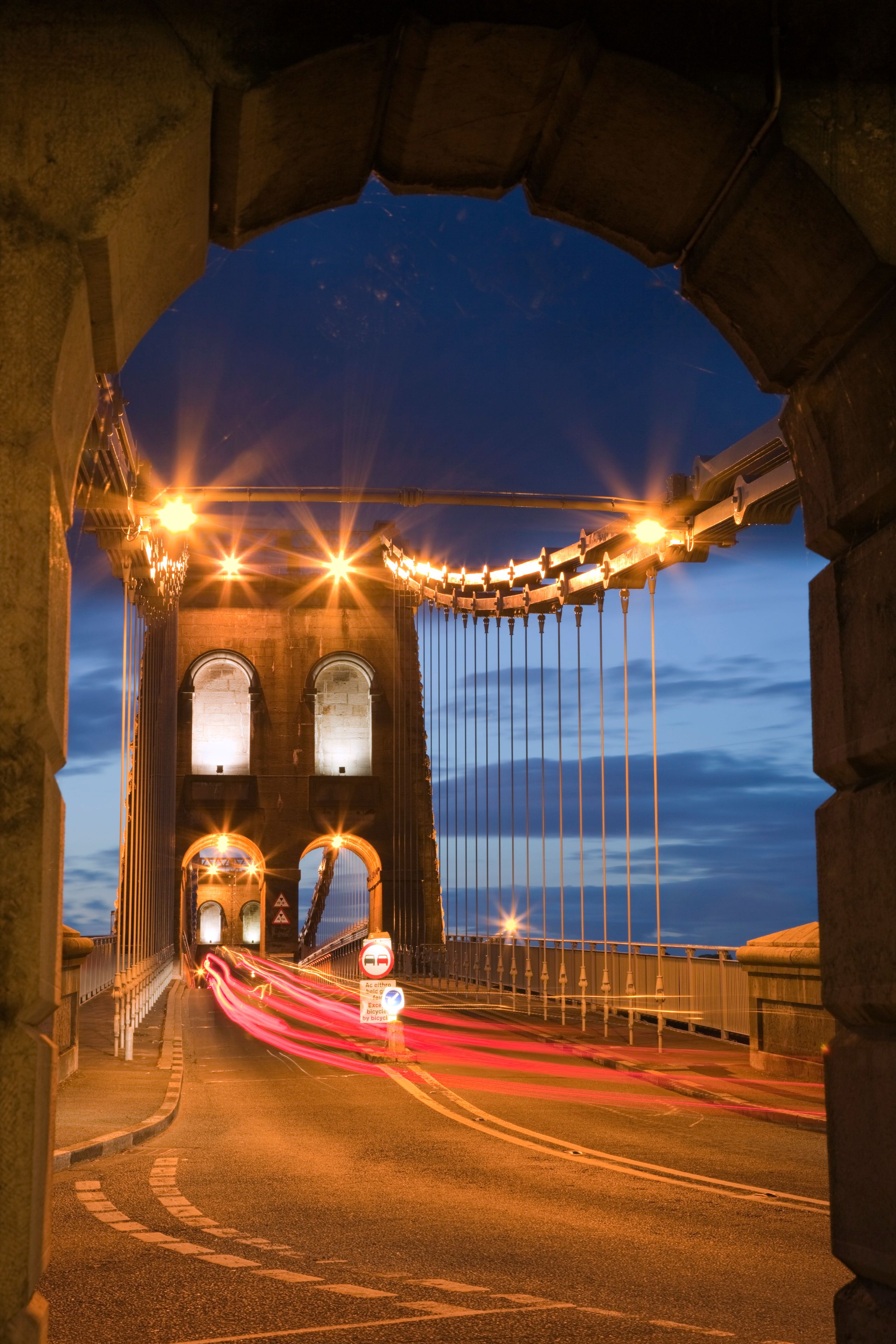 A bridge at dusk with illuminated lights and a red light trail from moving vehicles.