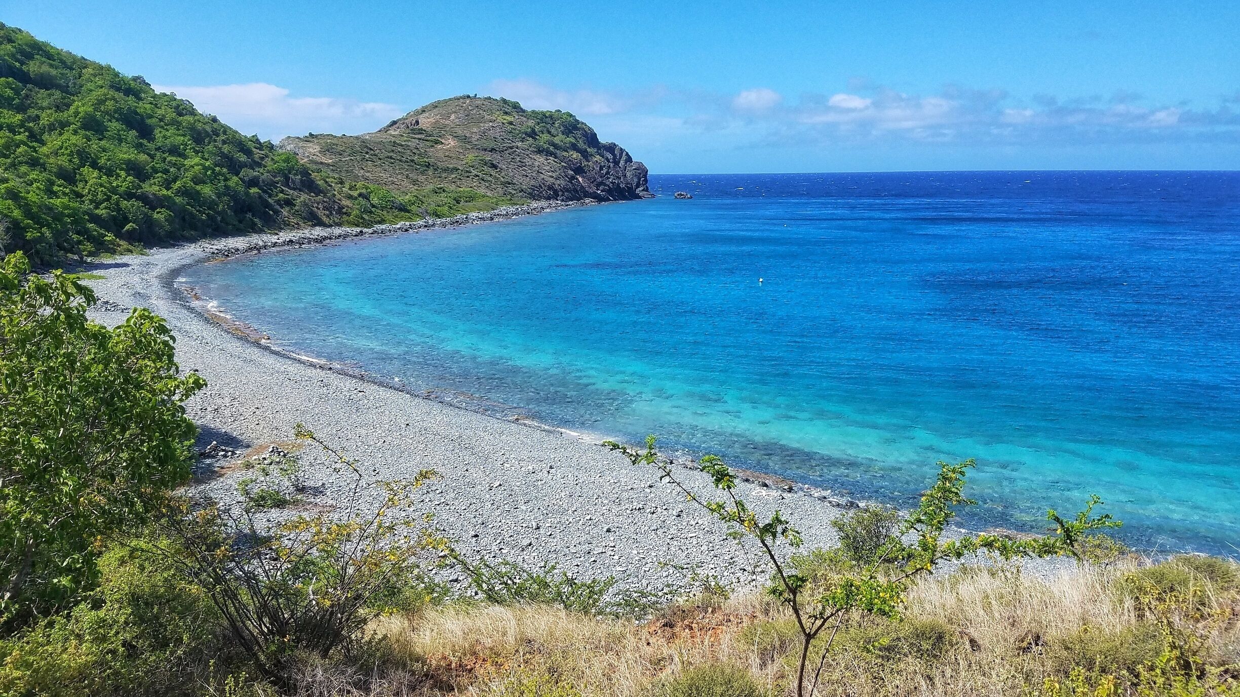 A coastal landscape with a pebble beach, clear turquoise waters, and a green hill in the background.