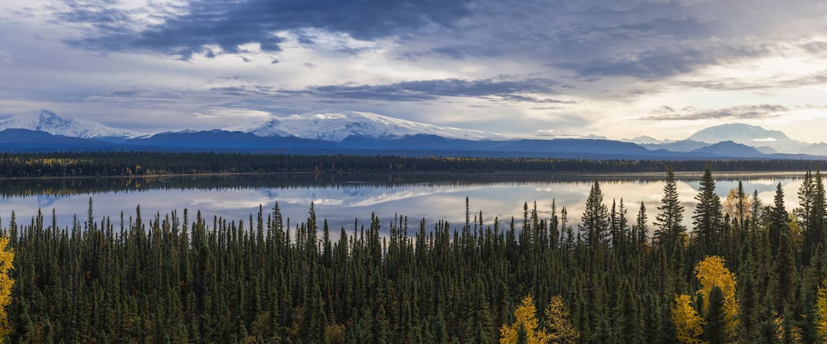 A lake surrounded by dense forest and snow-capped mountains under a cloudy sky.