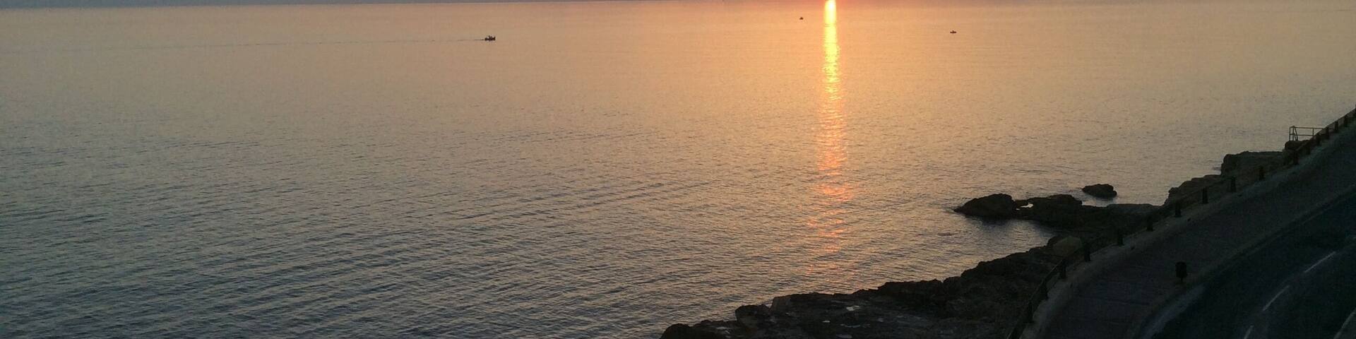 A sunset over a calm sea with rocky coastline and a road curving along the shore.