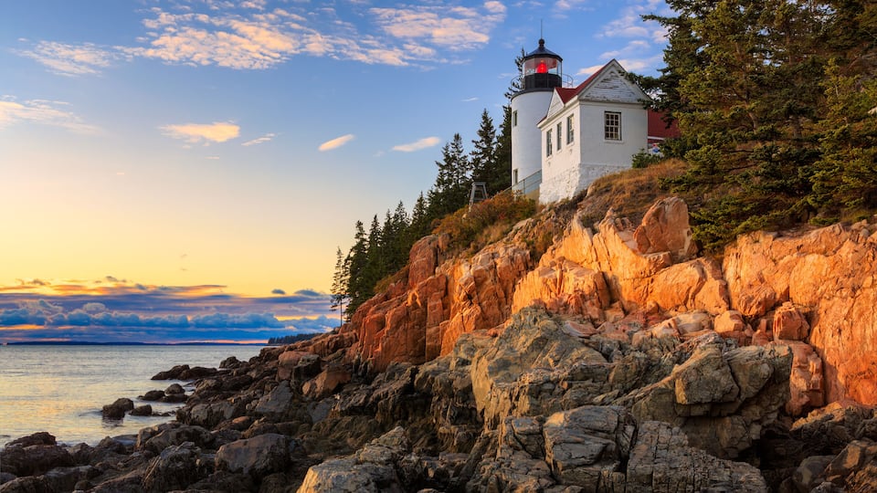 A lighthouse on a rocky cliff with a view of the ocean and a sunset sky.