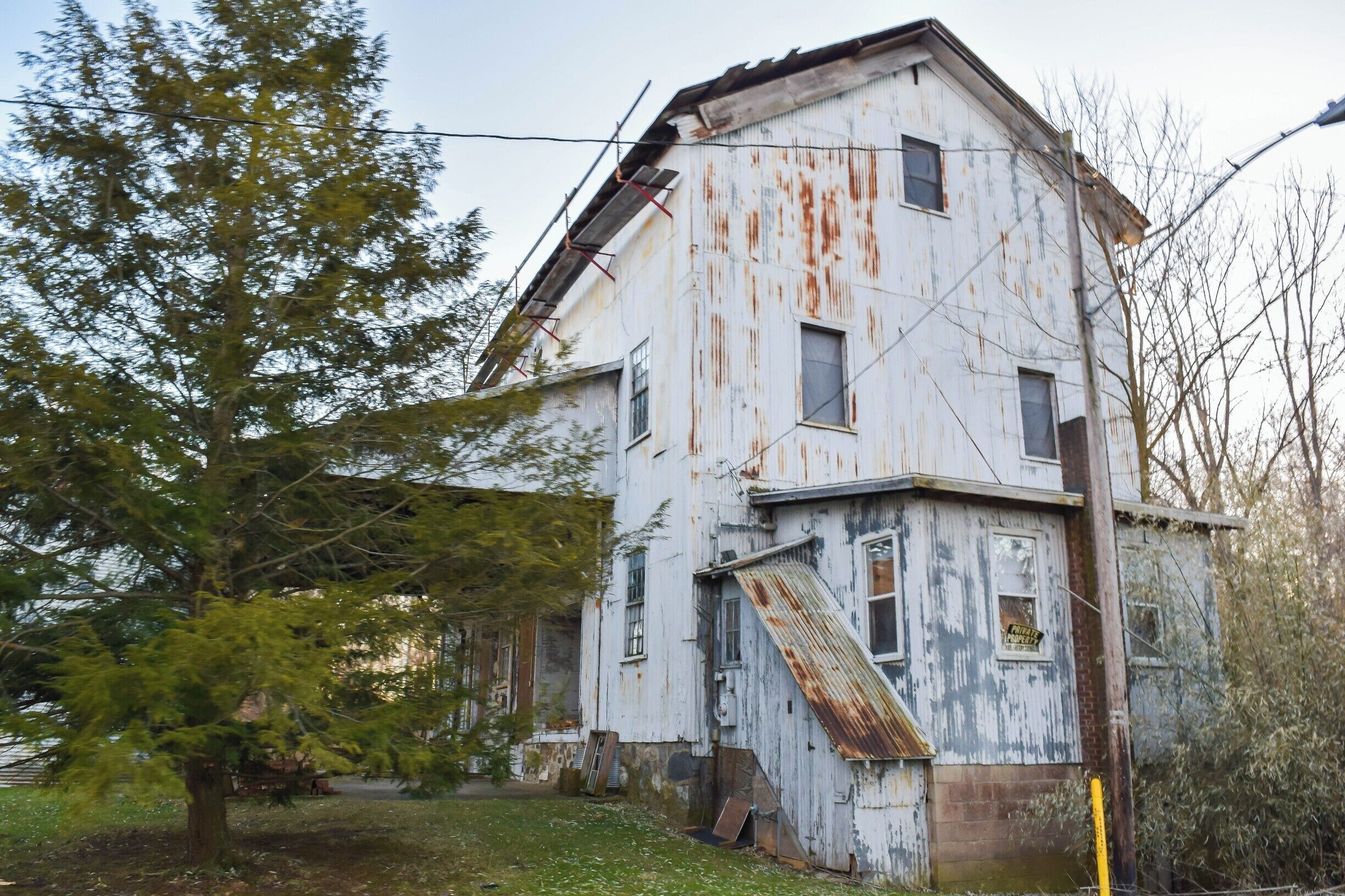A dilapidated, rusted building with a slanted roof, surrounded by trees and overgrown grass.