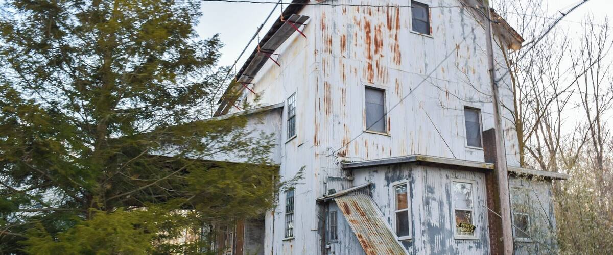 A dilapidated, rusted building with a slanted roof, surrounded by trees and overgrown grass.
