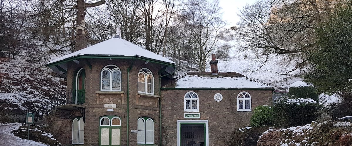Ein schneebedecktes Steinhaus mit grüner Tür und zahlreichen Fenstern, umgeben von Bäumen und einem verschneiten Weg.