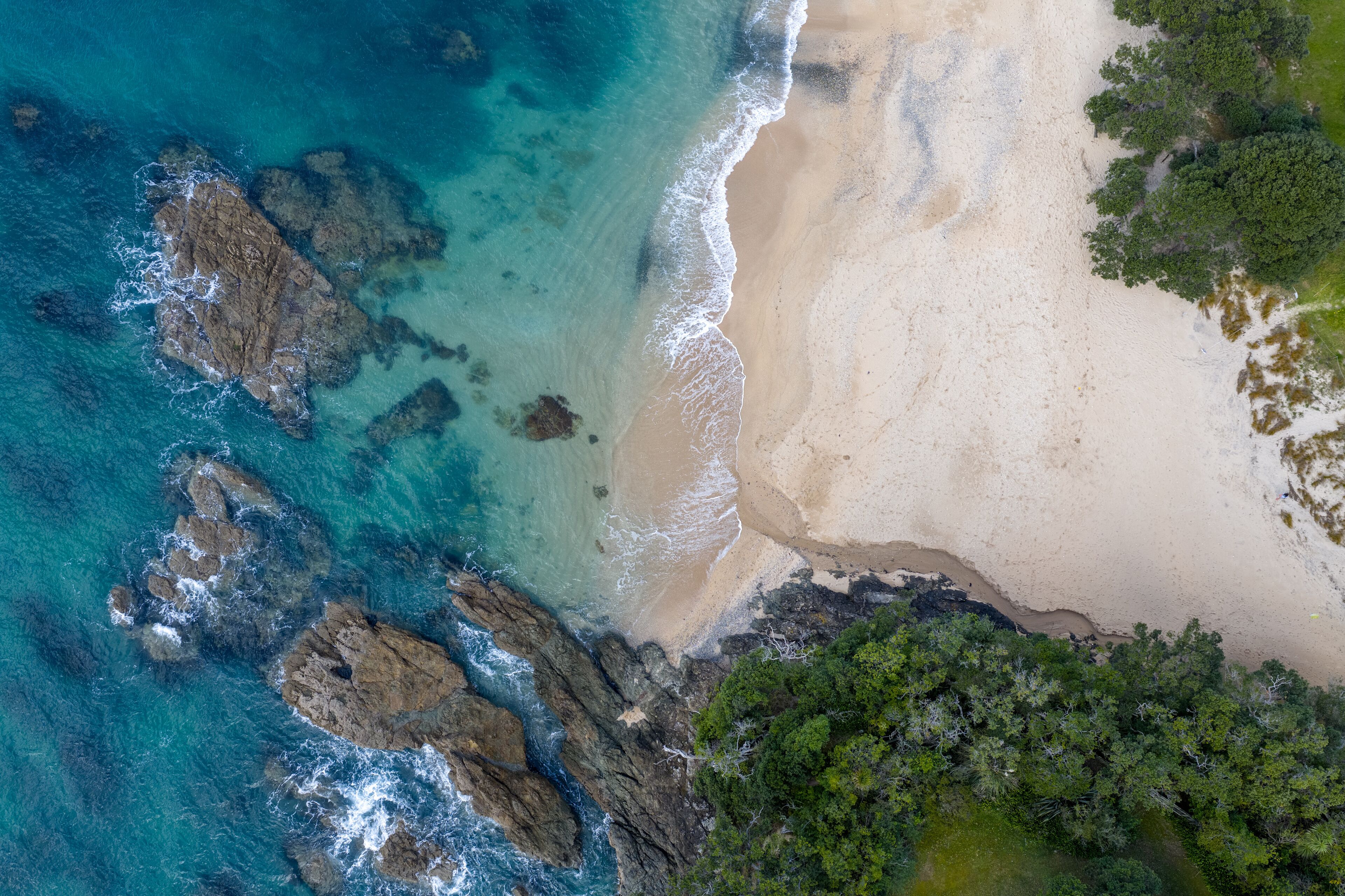 Aerial view of where the turquoise ocean meets the sandy shore near the rocks and vibrant green trees, Langs Beach, Northland Region, New Zealand.