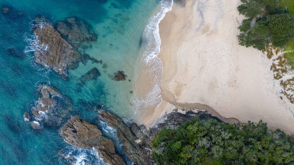 An aerial view of a beach with clear turquoise waters, rocky outcrops, and dense green foliage.