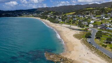 Aerial view of the coastline where azure waters meet the sandy beach next to a road lined with houses, Langs Beach, Northland Region, New Zealand.