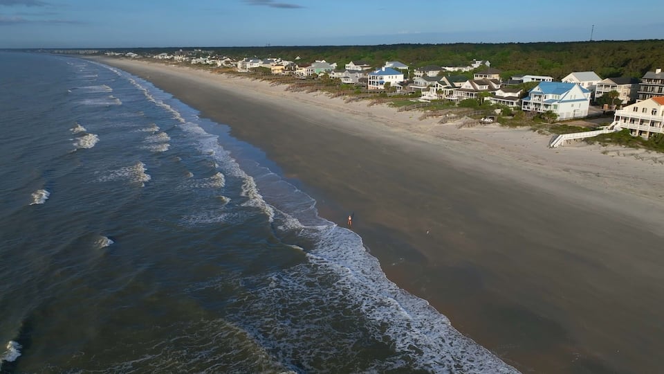 A beach with waves, a sandy shore, and houses along the coastline.