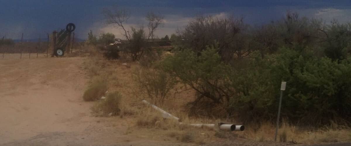 A dirt road leading to a metal structure in a desert landscape with sparse vegetation and a cloudy sky.
