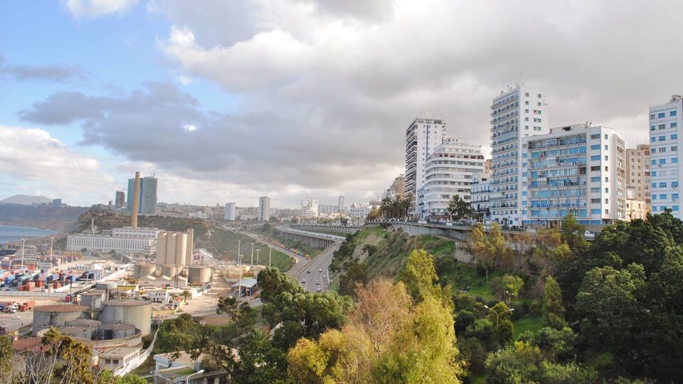 A coastal cityscape with modern high-rise buildings, a port area with shipping containers, and a hilly landscape with greenery.