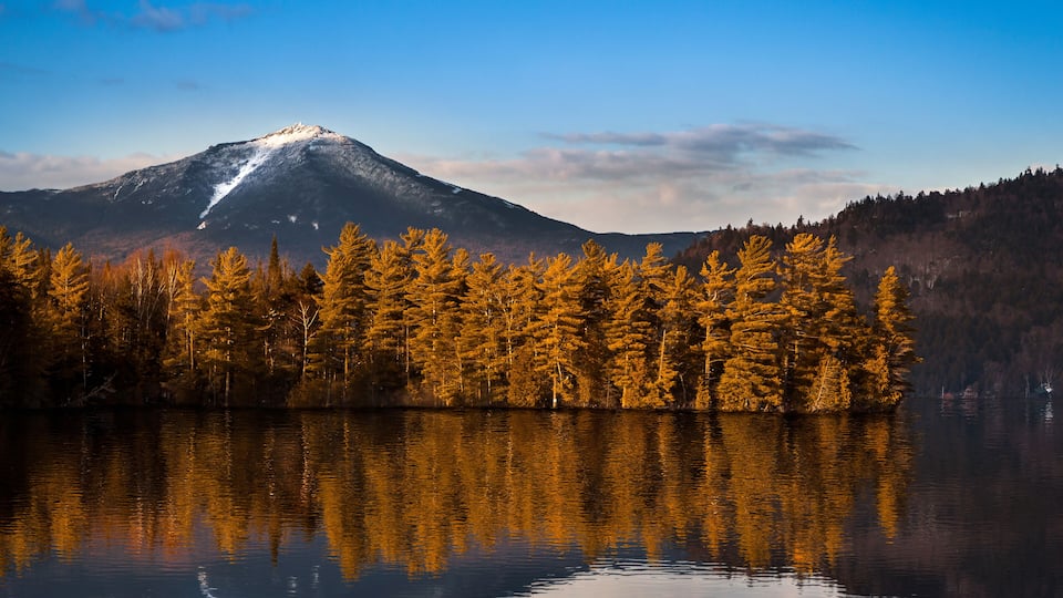A mountain with a snow-capped peak, a calm lake reflecting trees, and a clear blue sky.