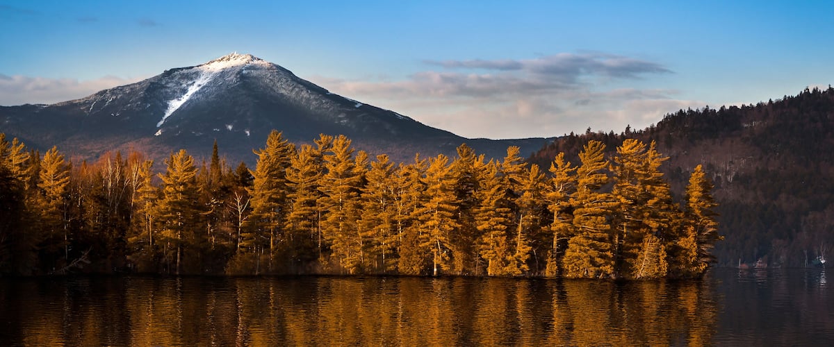A mountain with a snow-capped peak, a calm lake reflecting trees, and a clear blue sky.