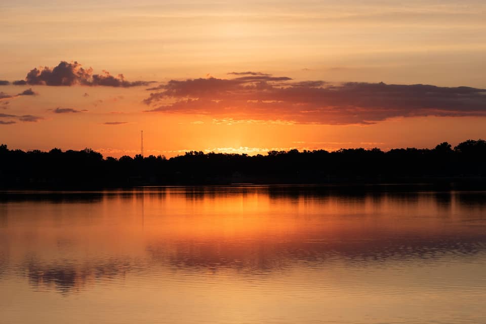 A sunset over a calm lake with a silhouette of trees and a distant tower.