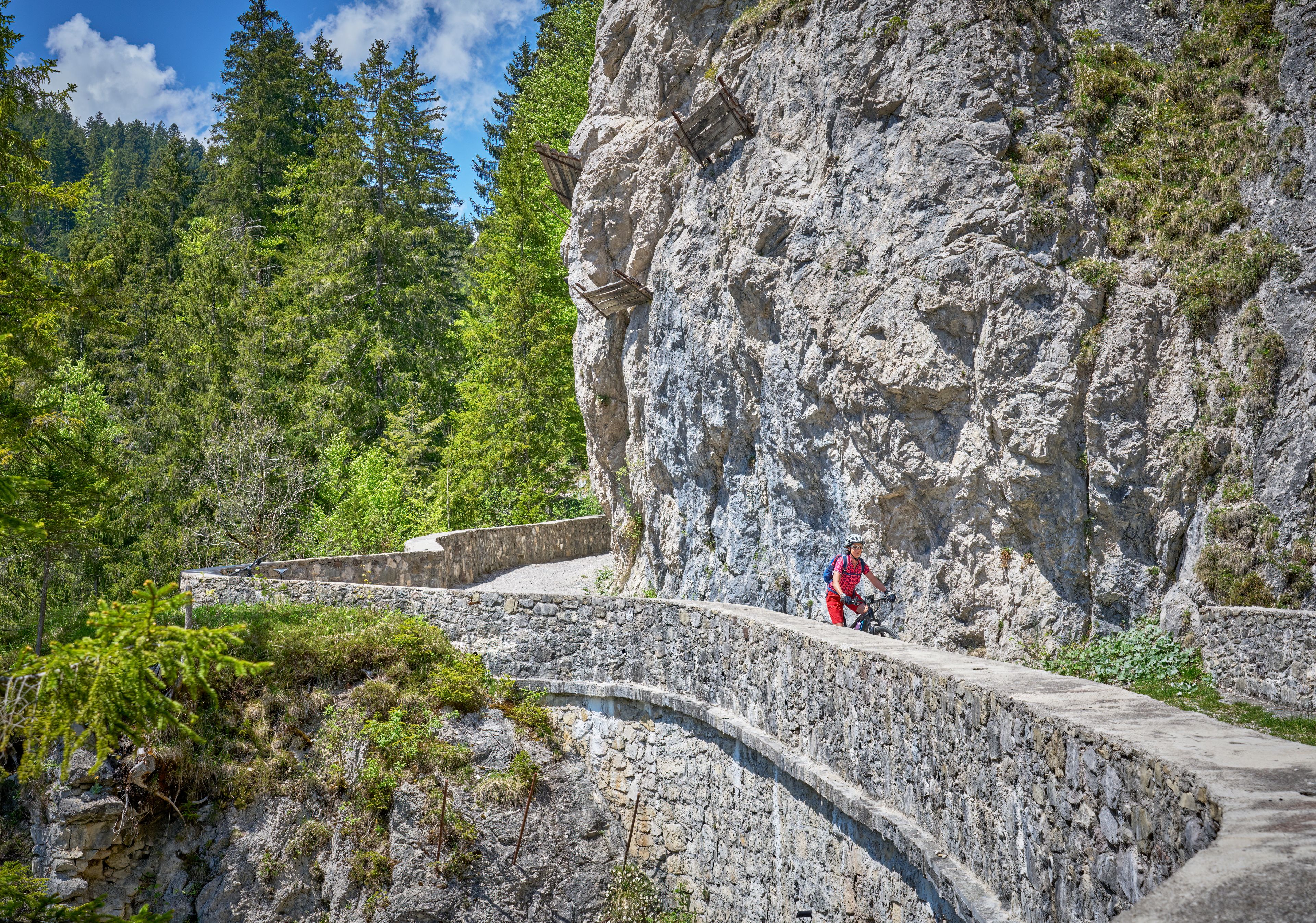 A mountain biker on a stone path with a rocky cliff and dense forest in the background.