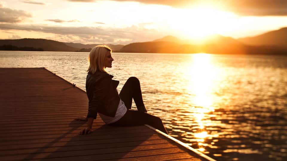 A person sitting on a wooden dock by a calm lake at sunset.