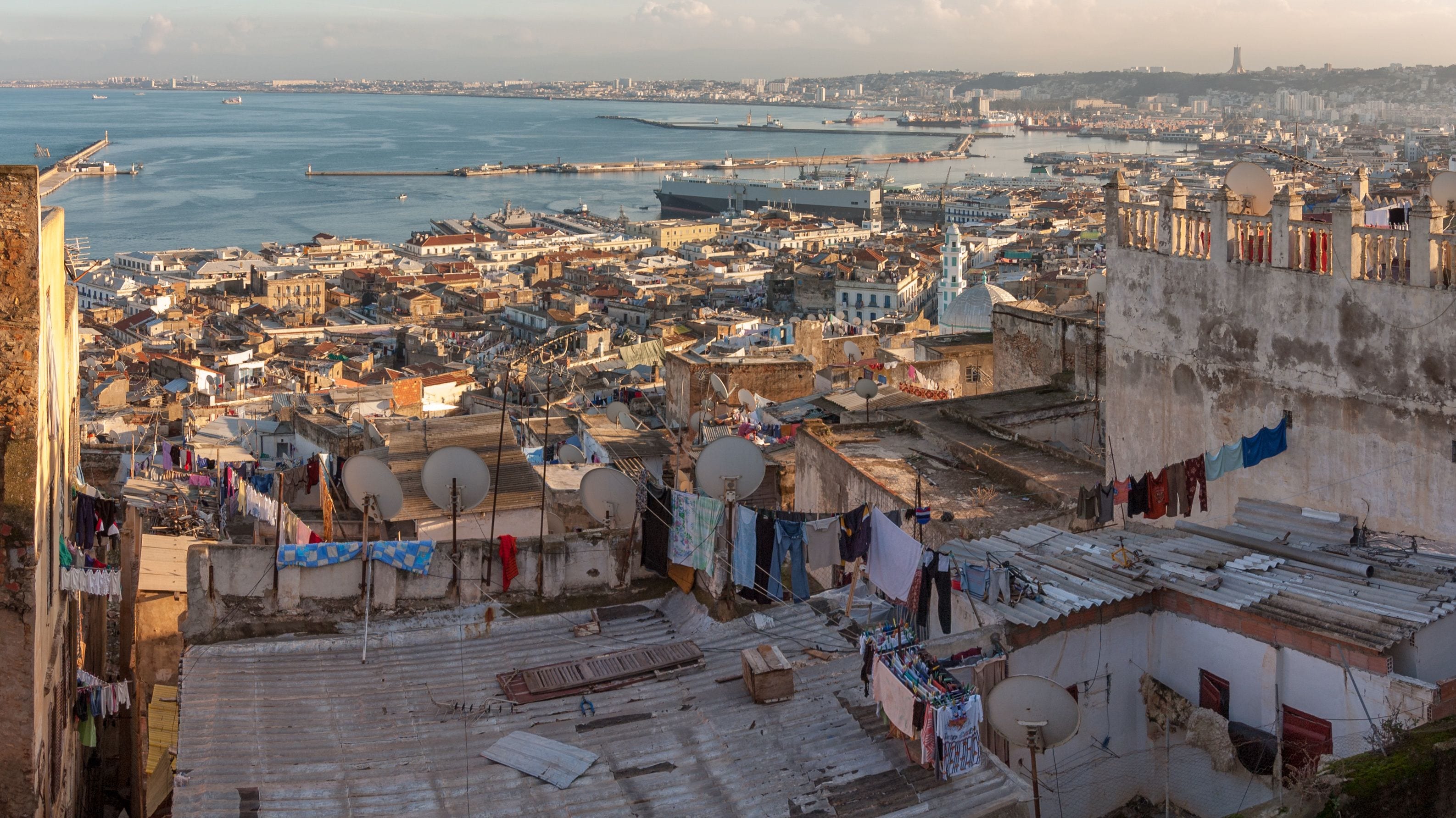 A coastal cityscape with a dense urban area, a harbor with ships, and a prominent tower in the distance.