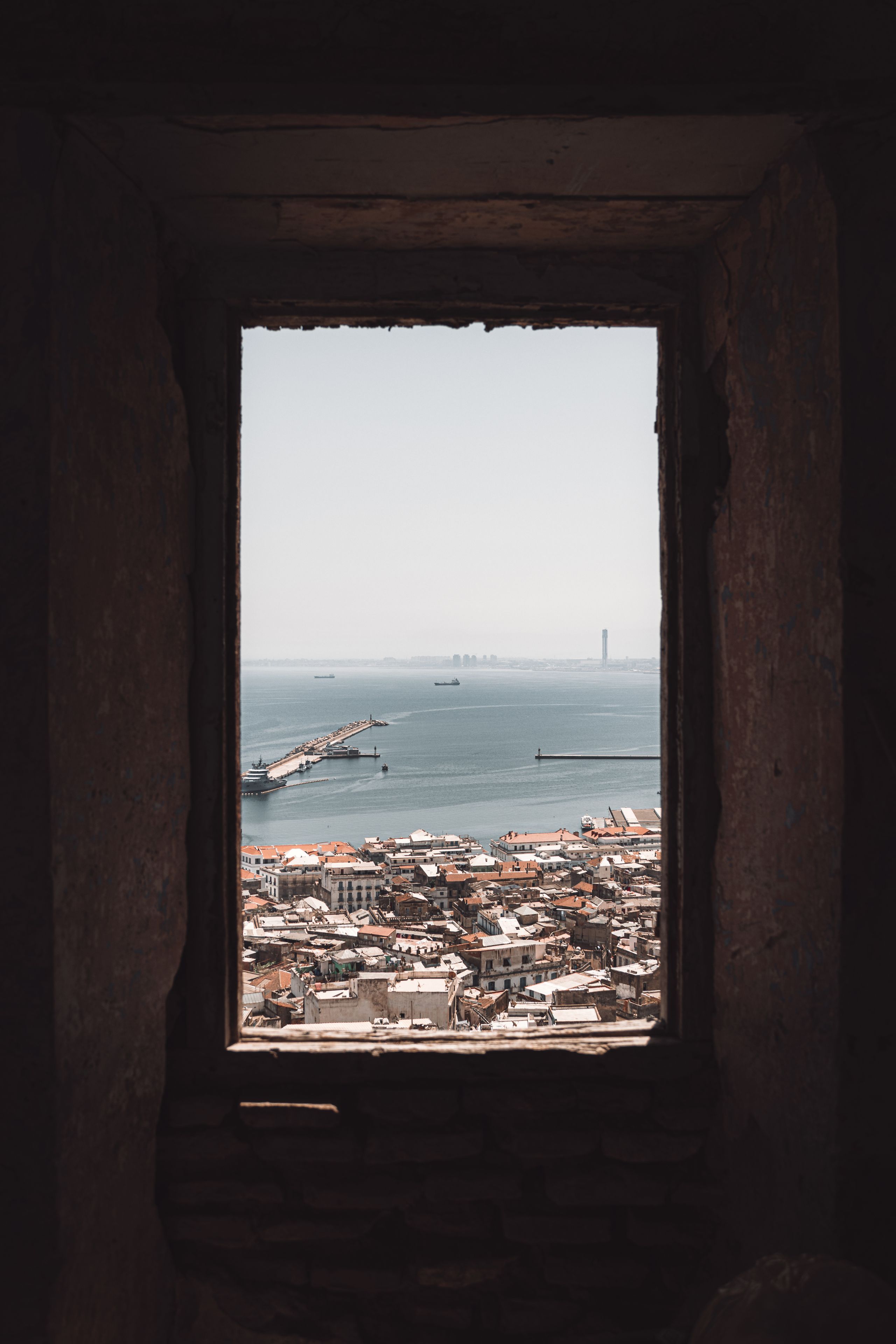 A coastal cityscape viewed through a window with a view of the sea, a pier, and distant buildings.