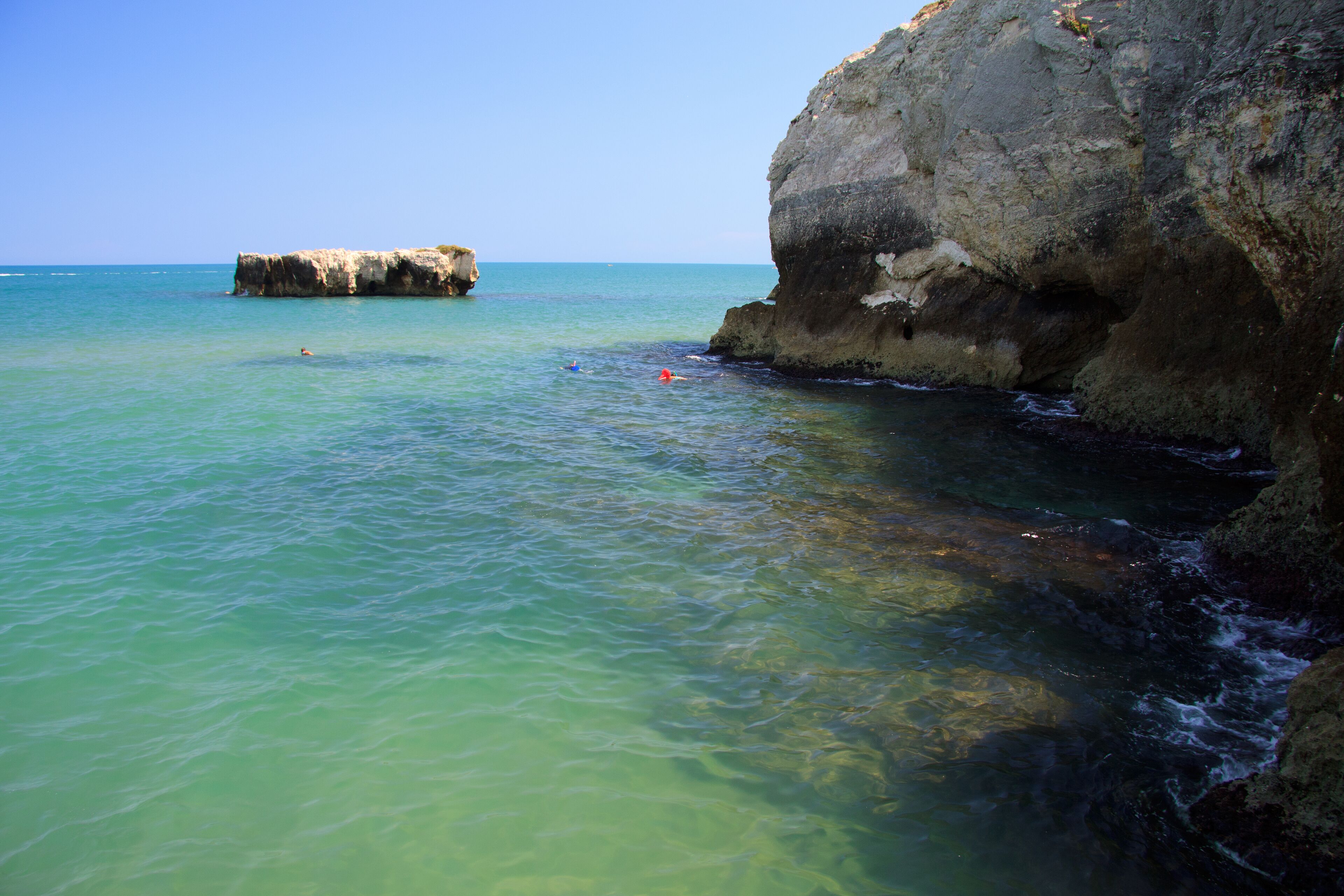 A coastal scene with clear turquoise water, a rocky cliff, and a small rock formation in the distance.