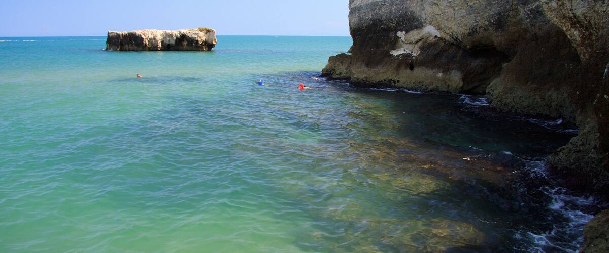A coastal scene with clear turquoise water, a rocky cliff, and a small rock formation in the distance.