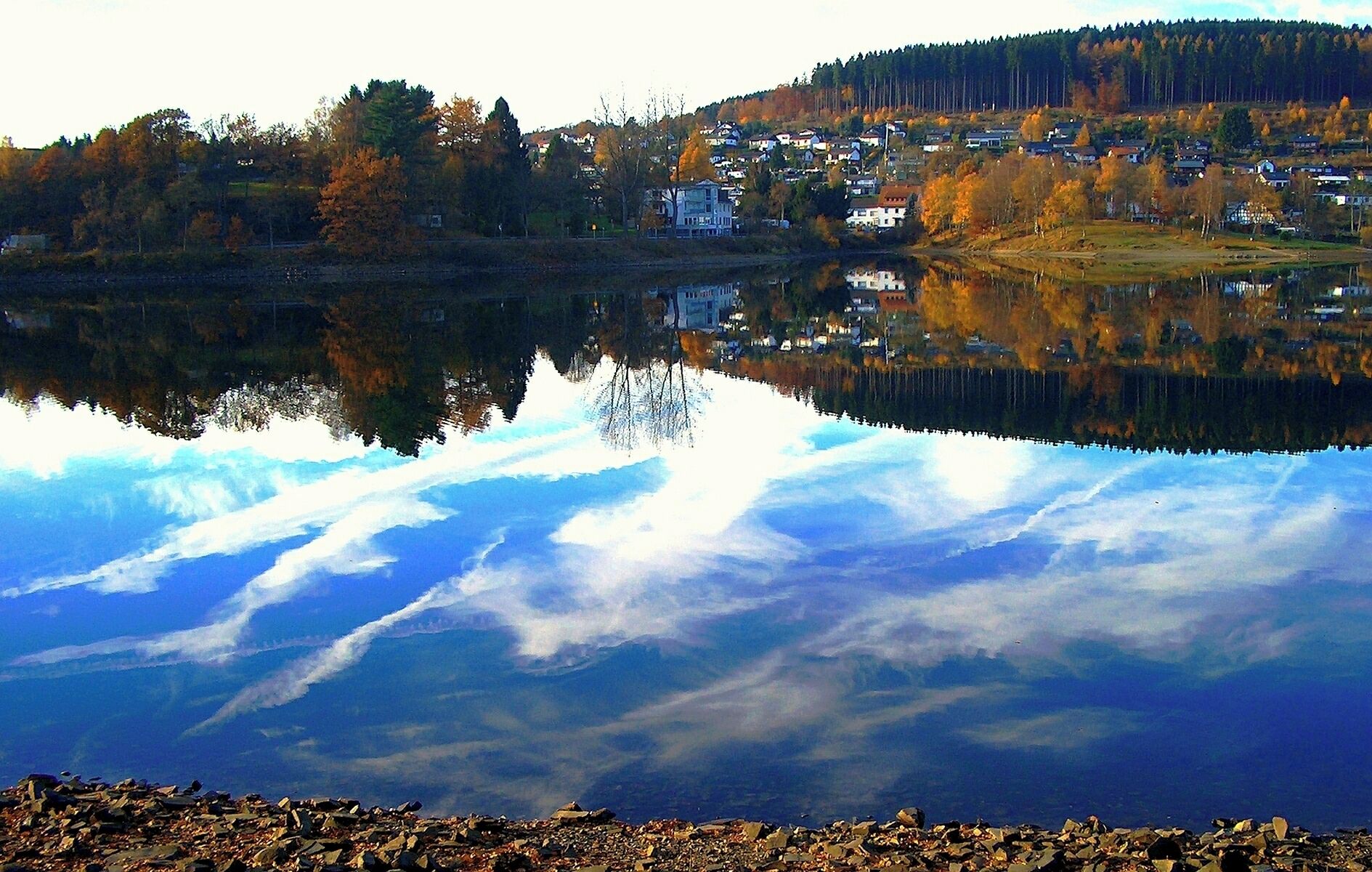 A calm lake reflecting a town with autumn-colored trees and a clear sky.