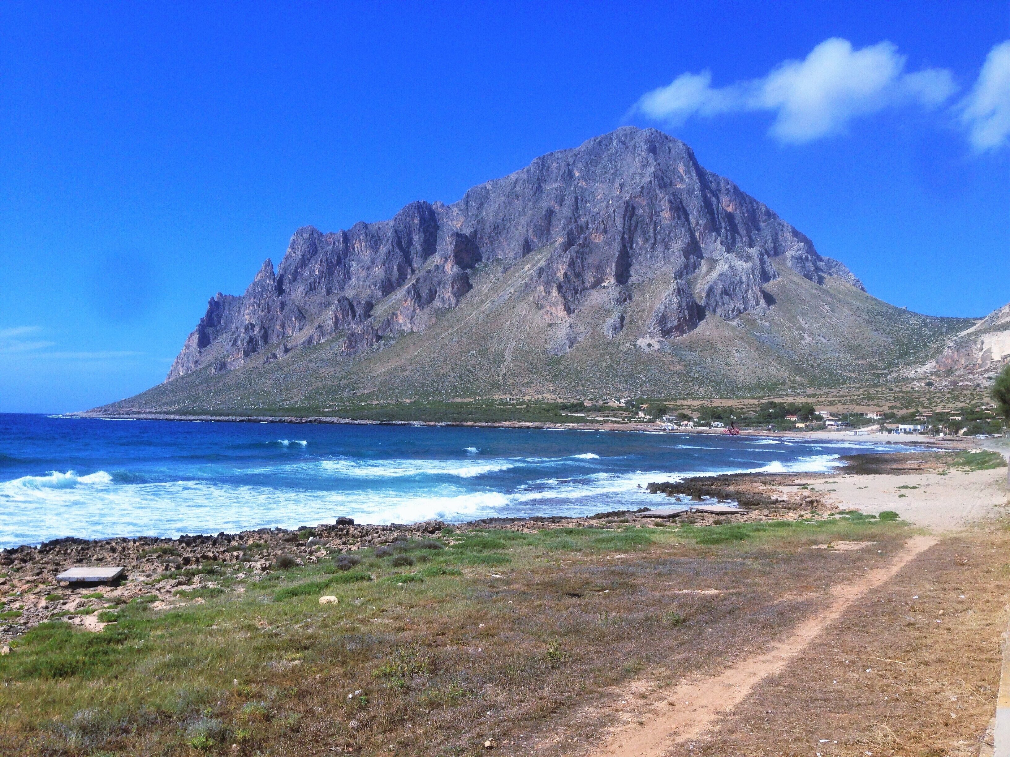 A rugged mountain with a clear blue sky, a beach with waves, and a dirt path leading to the shore.