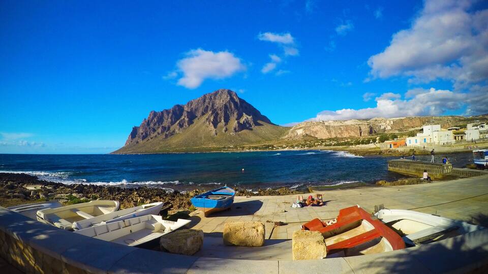 A coastal scene with a rocky shoreline, several boats, and a prominent mountain in the background.