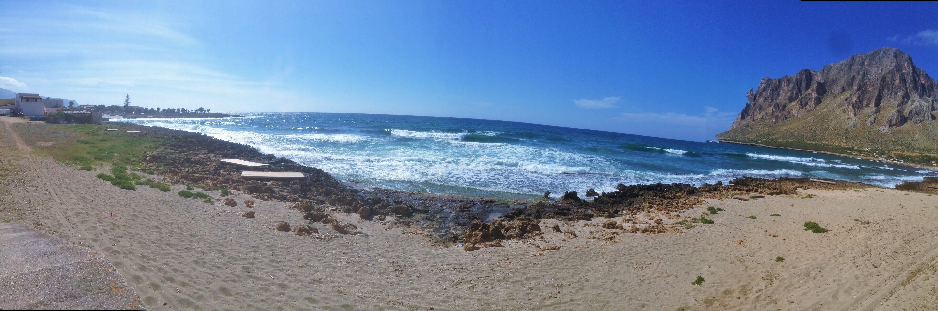 A beach with waves crashing onto rocky shorelines, a mountain in the background, and a clear blue sky.