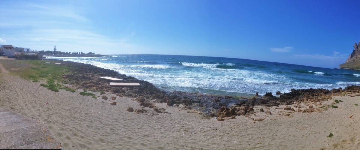 A beach with waves crashing onto rocky shorelines, a mountain in the background, and a clear blue sky.