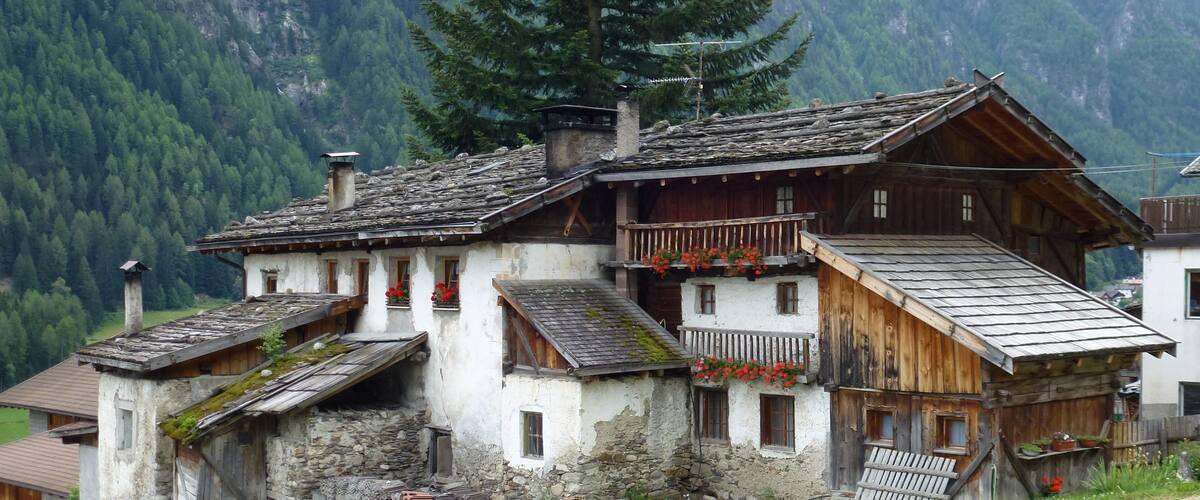 A traditional wooden house with a sloped roof, surrounded by lush greenery and mountains in the background.