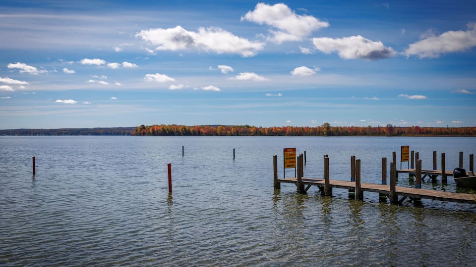 A calm lake with a wooden dock extending into the water, surrounded by autumn-colored trees and a clear blue sky with scattered clouds.