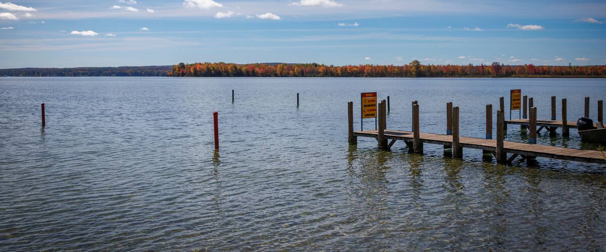 A calm lake with a wooden dock extending into the water, surrounded by autumn-colored trees and a clear blue sky with scattered clouds.