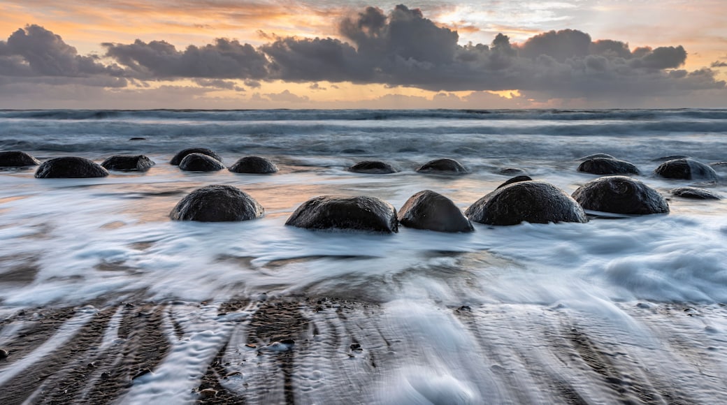 A beach with large rocks in the water and a sunset sky.