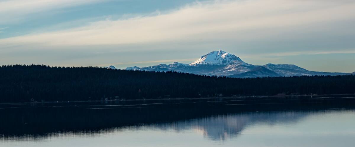 Dramatic image of mount Lassen reflecting in lake Almanor in Northern California with clear calm waters and cloudy skies.