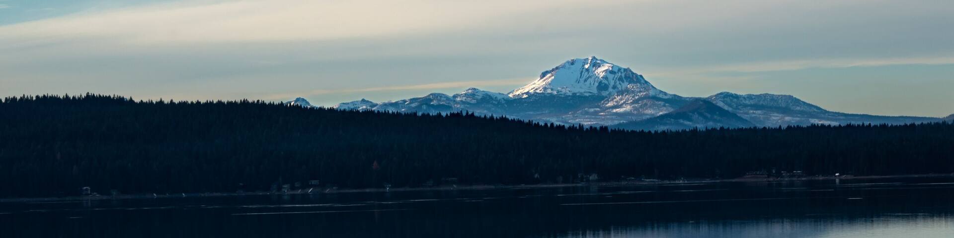 A snow-capped mountain, a calm lake, and a forested area under a clear sky.