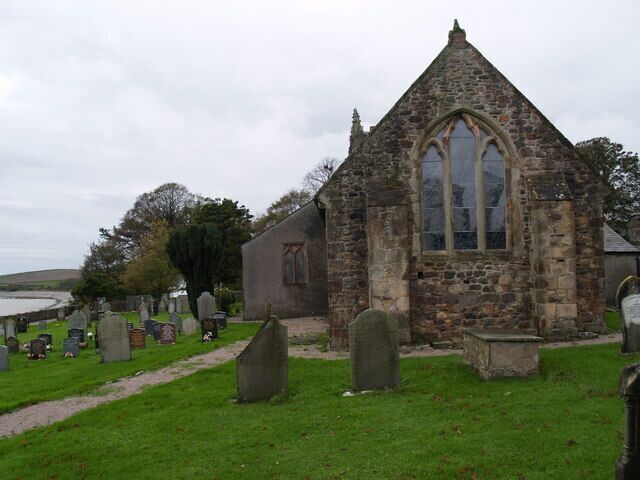 A stone church with a pointed roof and a large arched window, surrounded by a cemetery with headstones.
