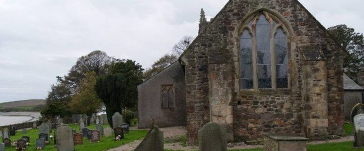 A stone church with a pointed roof and a large arched window, surrounded by a cemetery with headstones.