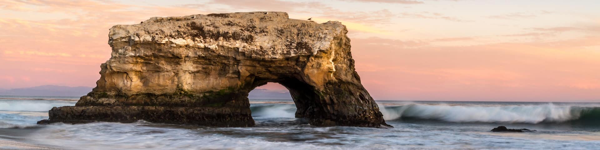 A natural rock arch formation on a beach with waves crashing below.